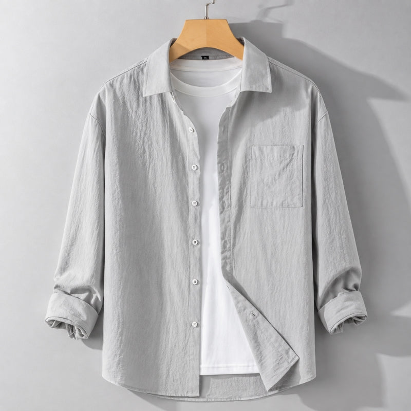 A light gray long-sleeve button-up shirt with a chest pocket, layered over a white t-shirt and hanging on a wooden hanger against a gray wall.