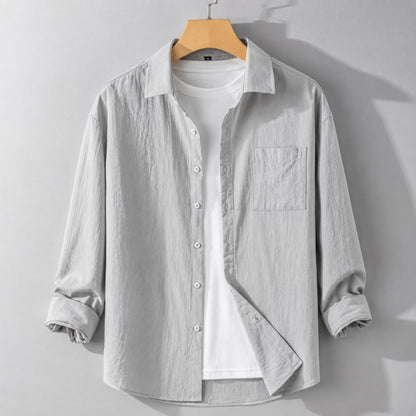A light gray long-sleeve button-up shirt with a chest pocket, layered over a white t-shirt and hanging on a wooden hanger against a gray wall.