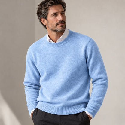 A man wearing a light blue crewneck knit sweater over a white collared shirt and dark trousers, standing against a neutral beige background.