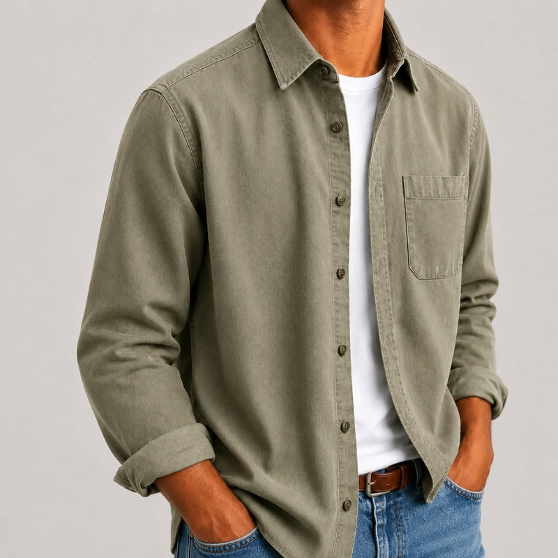 A man wearing an olive green long-sleeve button-up shirt over a white t-shirt and blue jeans, shown from the neck down against a light gray background.