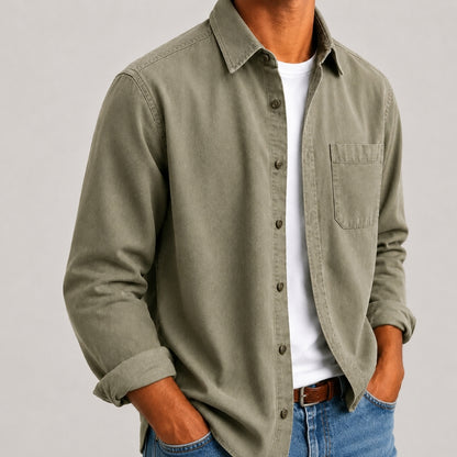 A man wearing an olive green long-sleeve button-up shirt over a white t-shirt and blue jeans, shown from the neck down against a light gray background.