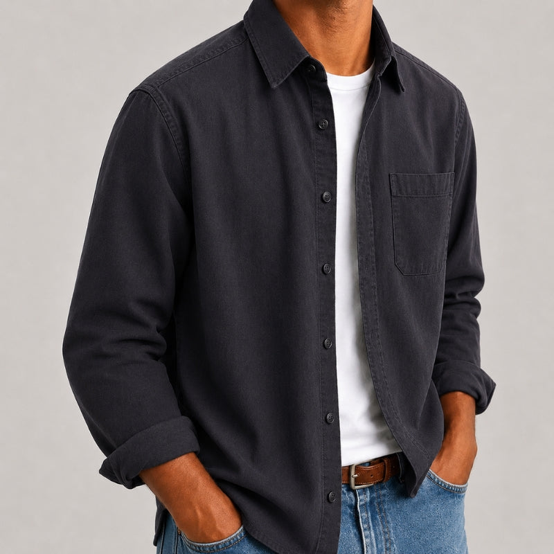 A man wearing a dark charcoal gray long-sleeve button-down shirt over a white t-shirt and blue jeans, shown from the neck down against a neutral gray background.
