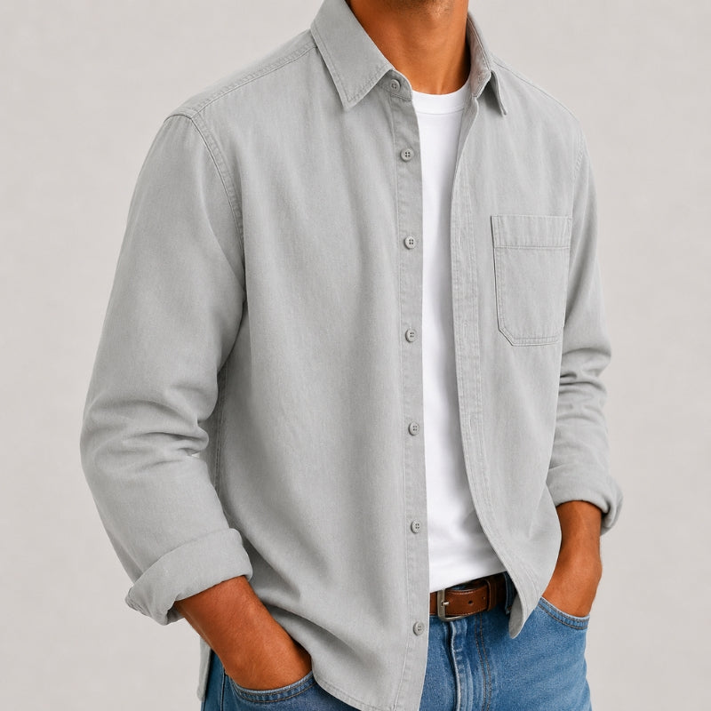 A man wearing a light gray long-sleeve button-down shirt over a white t-shirt and blue jeans, shown from the chest down against a neutral light gray background.