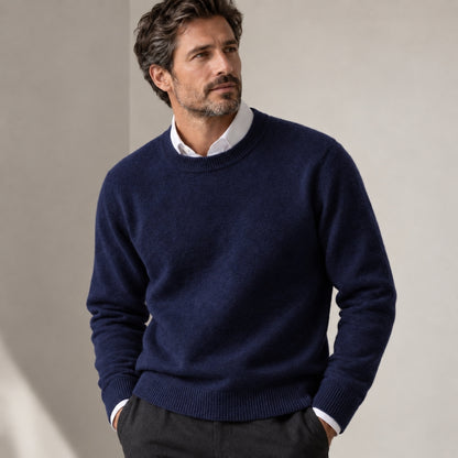 A man wearing a navy blue crewneck sweater over a white collared shirt and dark trousers, standing against a neutral beige wall.