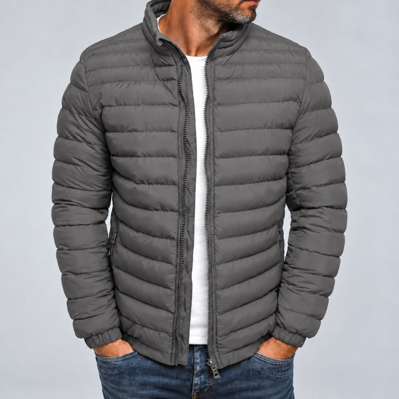 A man wearing a dark gray quilted puffer jacket over a white t-shirt and blue jeans, shown from the chin down against a light gray studio background.