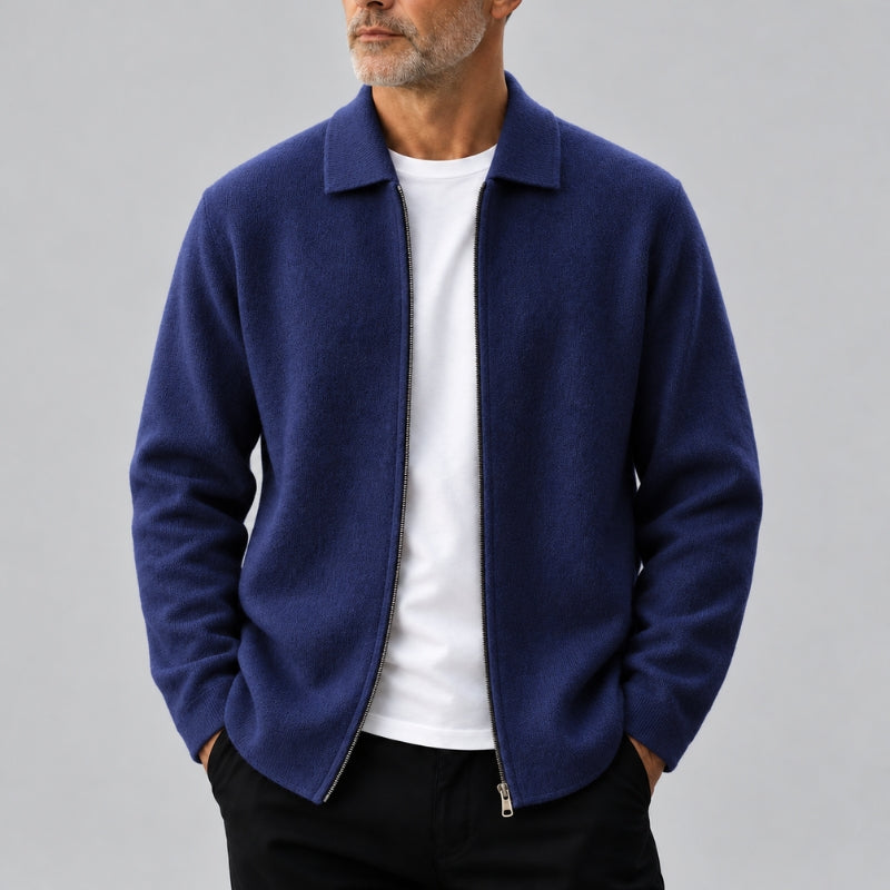 A man wearing a navy blue zip-up jacket over a white t-shirt and black pants, shown from the chin down against a solid gray background.
