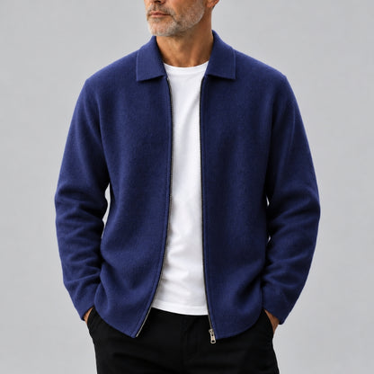 A man wearing a navy blue zip-up jacket over a white t-shirt and black pants, shown from the chin down against a solid gray background.