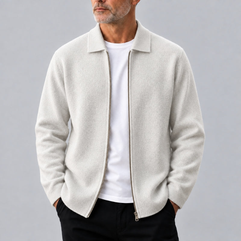 A man wearing a light beige zip-up jacket over a white t-shirt and black pants, standing against a solid gray background.