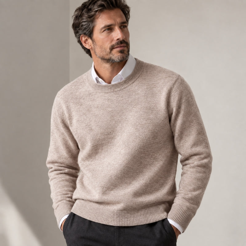 A man wearing a light beige crewneck knit sweater over a white collared shirt and dark trousers, standing against a neutral beige wall.