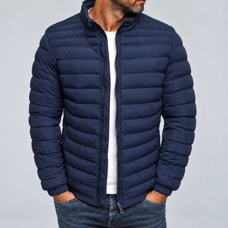 A man wearing a navy blue puffer jacket with horizontal quilting, a stand-up collar, and a front zipper, paired with a white t-shirt and blue jeans, against a plain gray background.