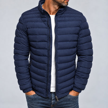 A man wearing a navy blue puffer jacket with horizontal quilting, a stand-up collar, and a front zipper, paired with a white t-shirt and blue jeans, against a plain gray background.
