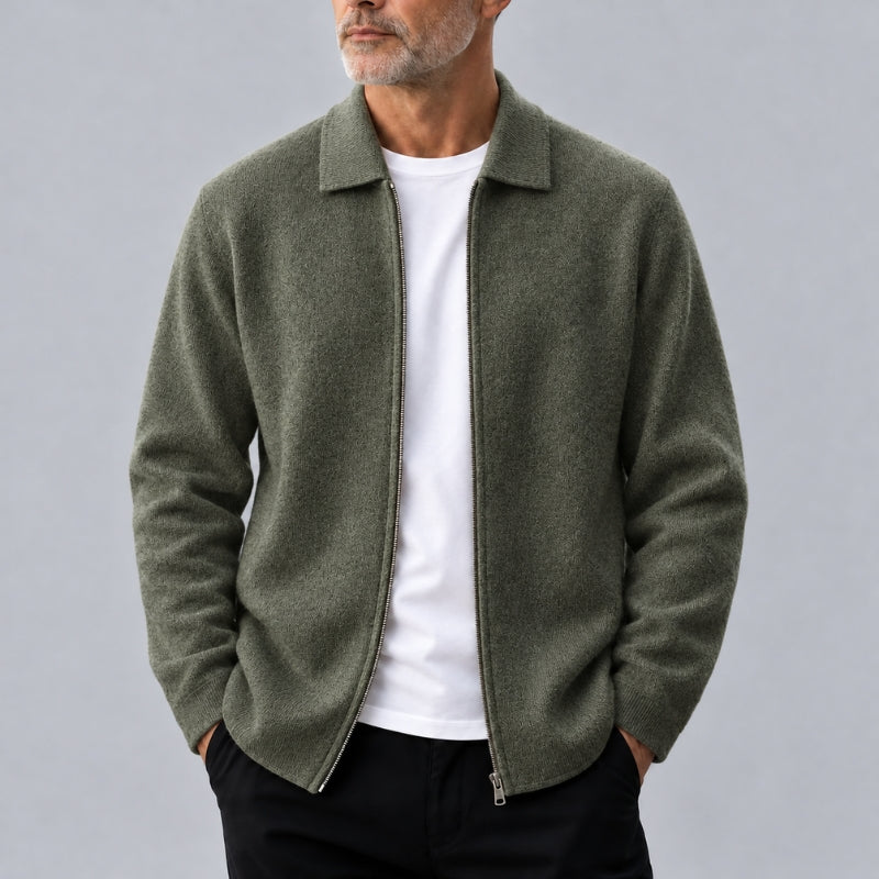 A man wearing an olive green zip-up collar jacket over a white t-shirt and black pants, photographed against a neutral gray background.
