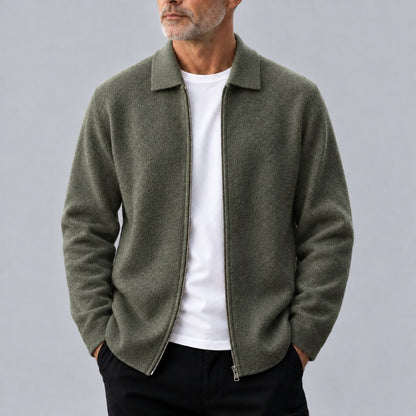 A man wearing an olive green zip-up collar jacket over a white t-shirt and black pants, photographed against a neutral gray background.