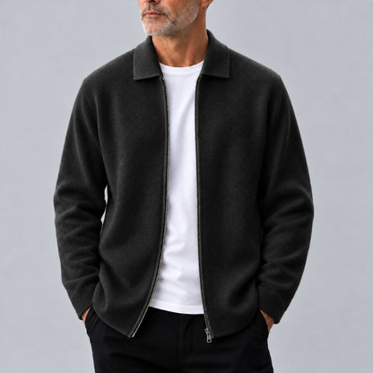 A man wearing a dark charcoal gray zip-up jacket over a white t-shirt and black pants, standing against a solid gray studio background.