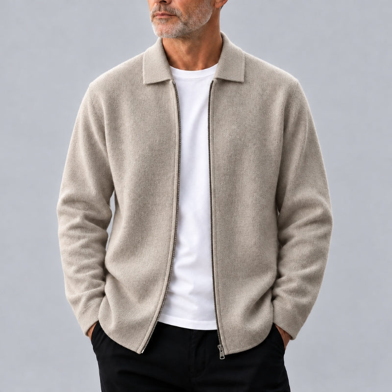 A man wearing a light beige zip-up knit jacket over a white t-shirt and black pants, standing against a solid gray studio background.