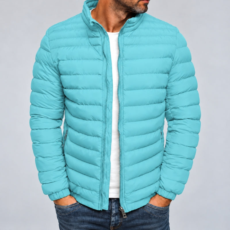 A man wearing a light blue-green puffer jacket over a white t-shirt and dark blue jeans, shown from the chin down against a plain gray studio background.