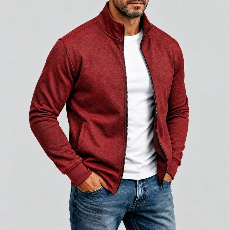 A man wearing a heathered red zip-up jacket over a white t-shirt and blue jeans, standing against a light gray studio background.