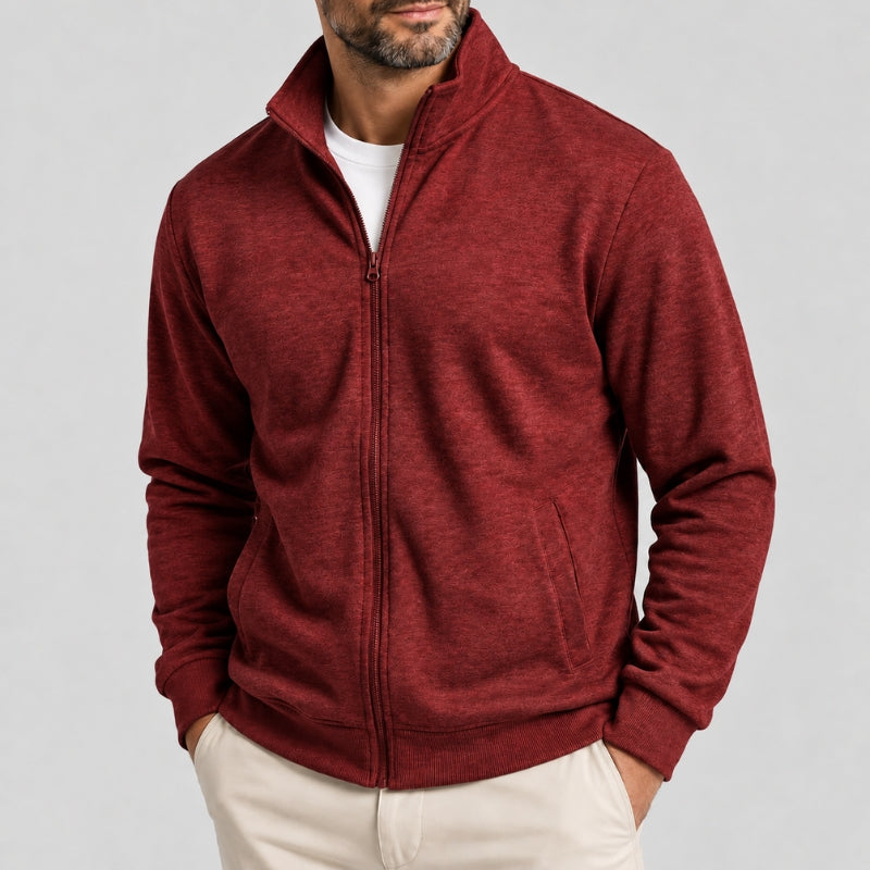 A man wearing a heathered burgundy zip-up fleece jacket over a white t-shirt and cream pants, photographed against a light gray studio background.