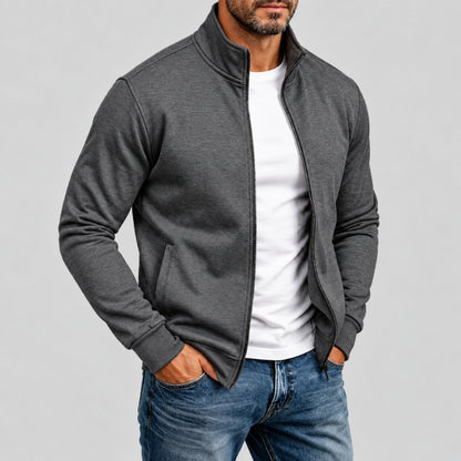 A man wearing a dark gray zip-up fleece jacket over a white t-shirt and blue jeans, standing against a solid gray background.