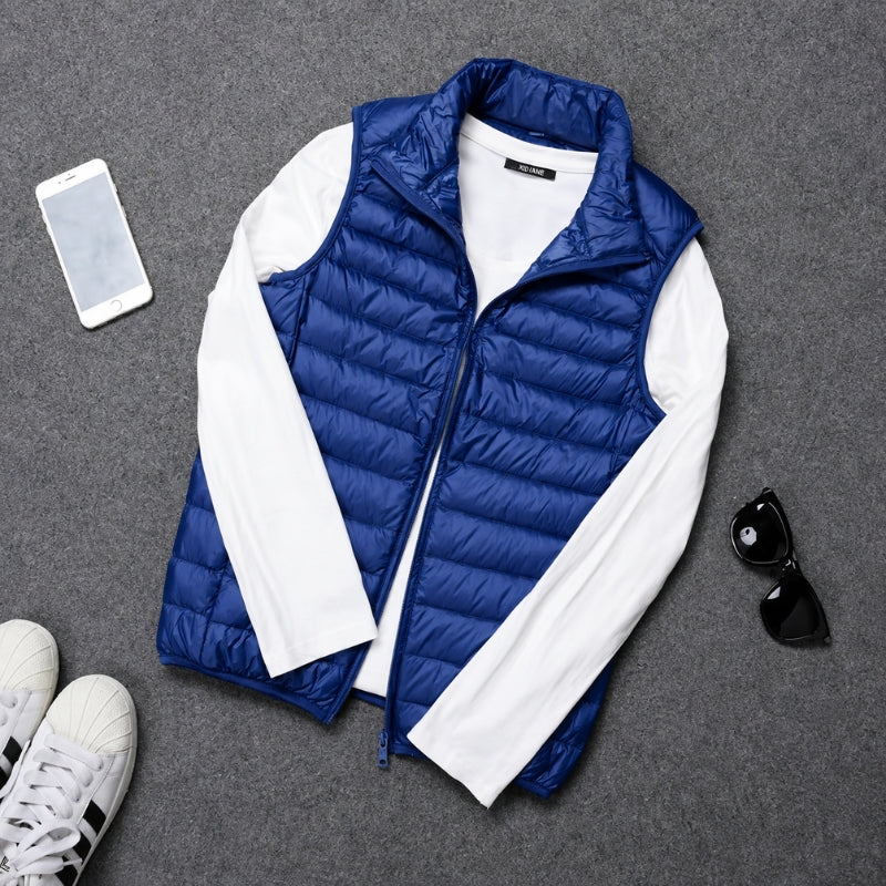 A blue puffer vest layered over a white long-sleeve shirt, arranged in a flat lay on a gray textured surface with a smartphone, sunglasses, and sneakers nearby.