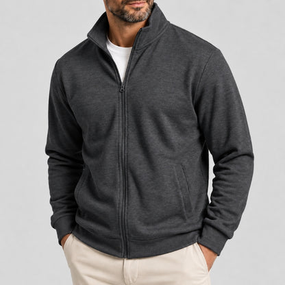 A man wearing a dark gray heather zip-up fleece jacket over a white t-shirt and beige pants, shown from the chin down against a light gray studio background.
