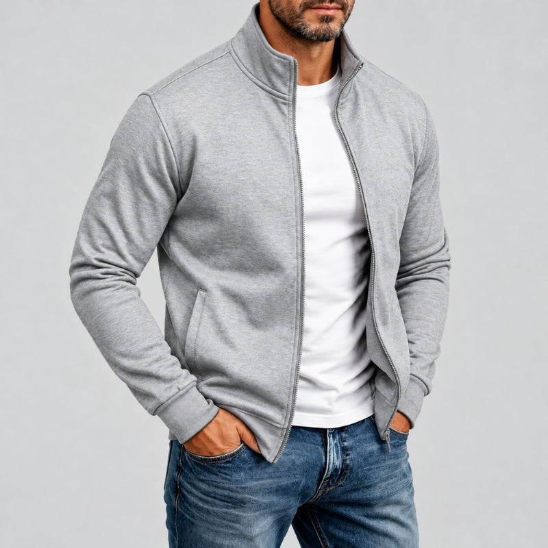 A man wearing a light gray zip-up fleece jacket over a white t-shirt and blue jeans, standing against a solid gray studio background.