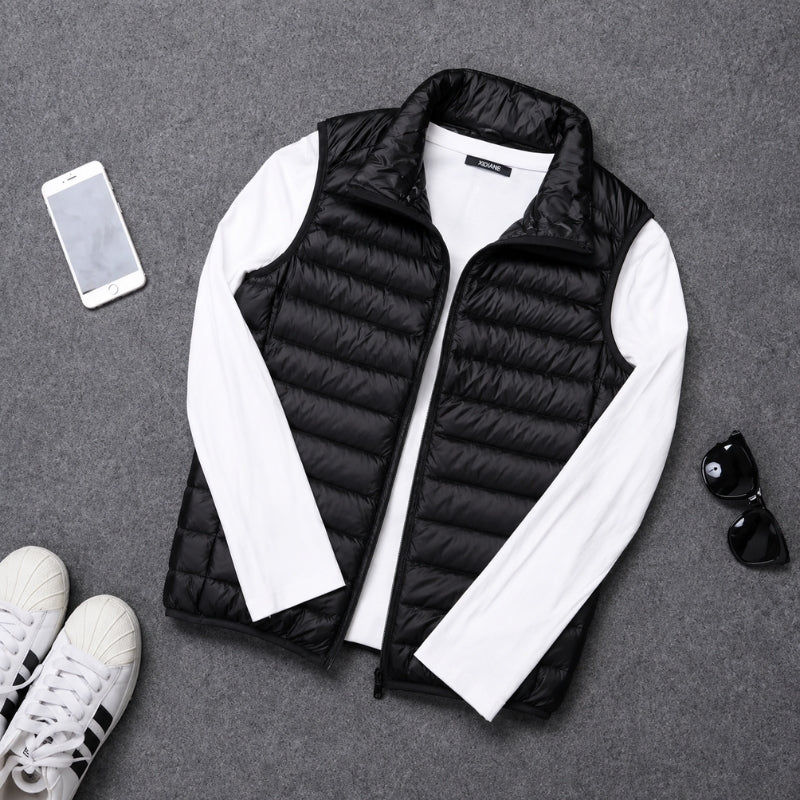 A black quilted puffer vest styled in a flat lay with a white long-sleeve shirt underneath, accompanied by a smartphone, sunglasses, and sneakers on a gray textured background.