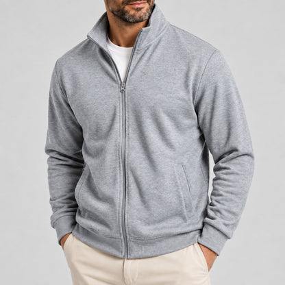 A man wearing a light gray heathered zip-up fleece jacket with a stand collar, paired with a white t-shirt and beige trousers, against a solid light gray background.