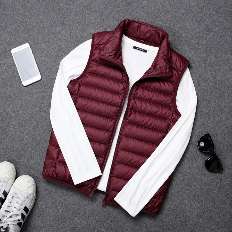 A burgundy puffer vest layered over a white long-sleeve shirt, styled in a flat lay with a smartphone, sunglasses, and sneakers on a gray textured background.