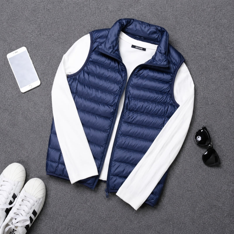 A navy blue puffer vest with white long sleeves, styled in a flat lay on a gray textured background with a smartphone, sunglasses, and white sneakers.