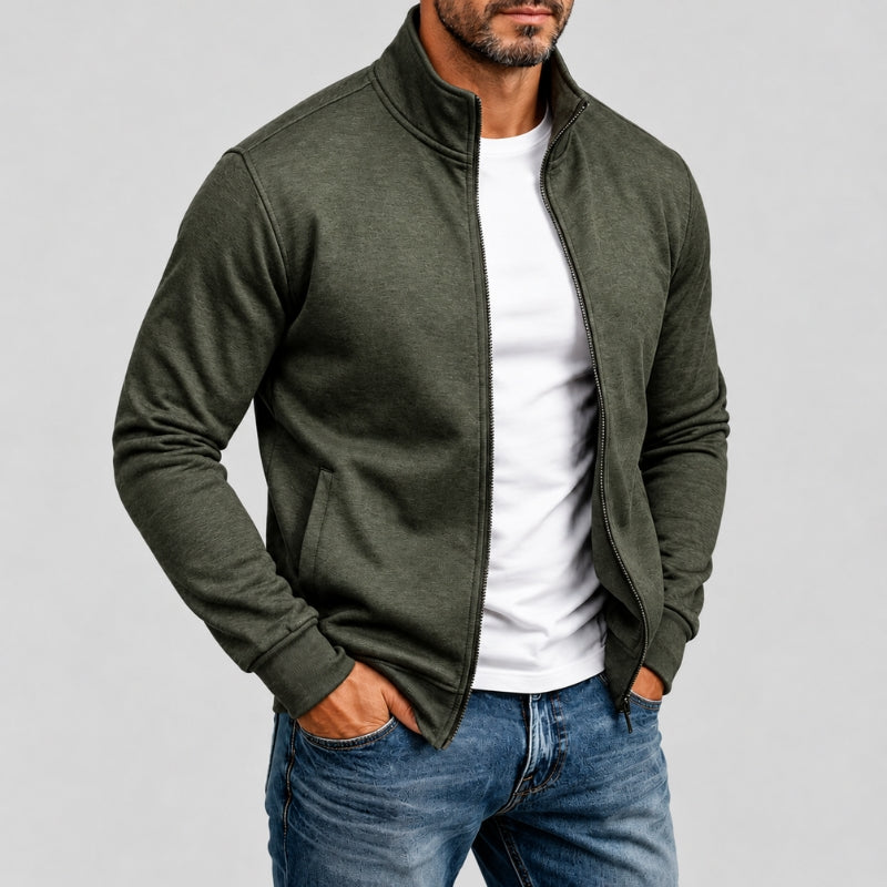 A man wearing a heathered olive green zip-up jacket over a white t-shirt and blue jeans, standing against a light gray studio background.