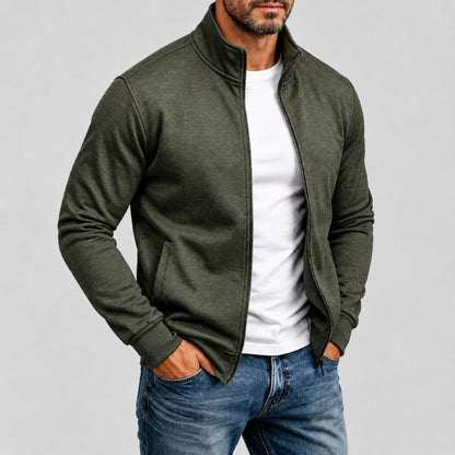 A man wearing a heathered olive green zip-up jacket over a white t-shirt and blue jeans, standing against a light gray studio background.