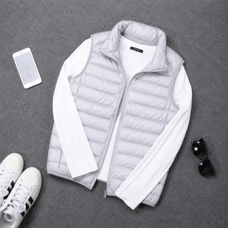 A light gray puffer vest layered over a white long-sleeve shirt, arranged in a flat lay on a gray textured surface with a smartphone, white sneakers, and black sunglasses nearby.