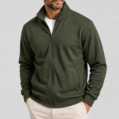 A man wearing an olive green full-zip fleece jacket over a white t-shirt and cream pants, shown from the chin down against a light gray studio background.