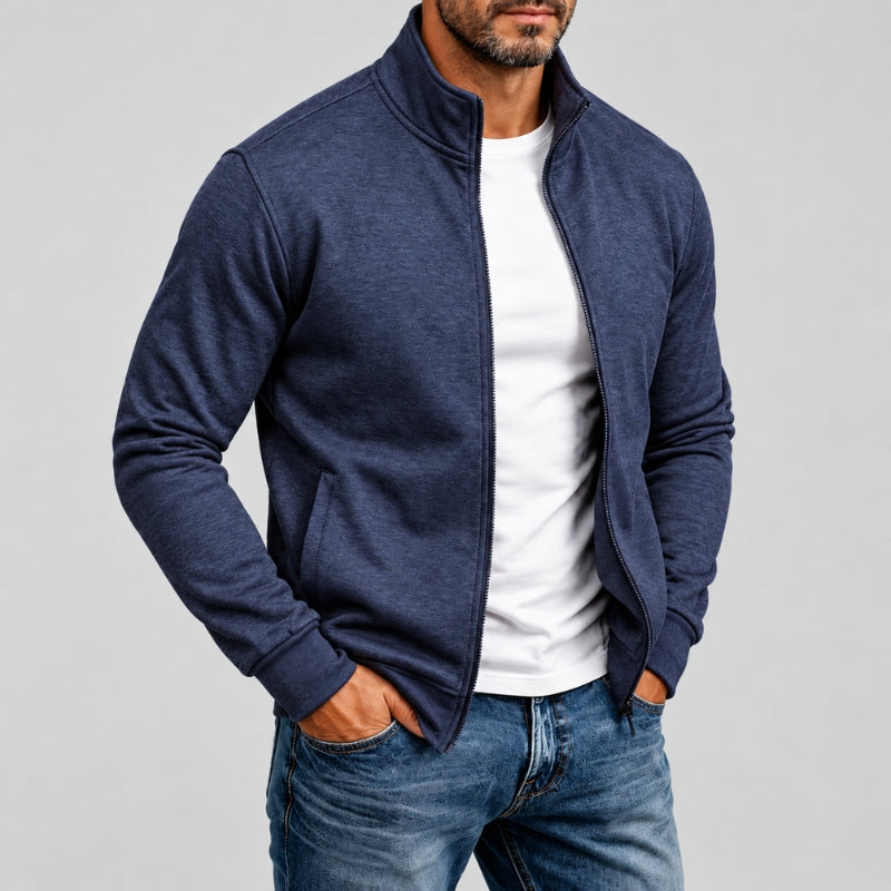 A man wearing a navy blue zip-up jacket over a white t-shirt and blue jeans, standing against a light gray studio background.
