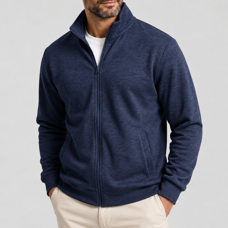 A man wearing a navy blue heathered zip-up fleece jacket over a white t-shirt and beige pants, shown from the chin down against a light gray studio background.