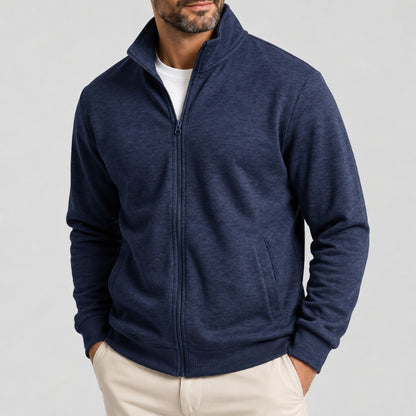 A man wearing a navy blue heathered zip-up fleece jacket over a white t-shirt and beige pants, shown from the chin down against a light gray studio background.