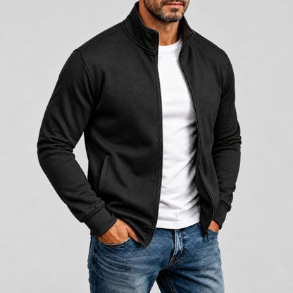 A man wearing a black zip-up jacket over a white t-shirt and blue jeans, standing against a light gray studio background.