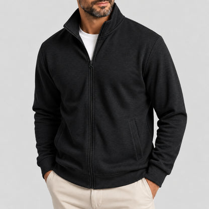 A man wearing a black zip-up fleece jacket with a stand-up collar, layered over a white t-shirt and paired with beige trousers, photographed against a solid light gray background.