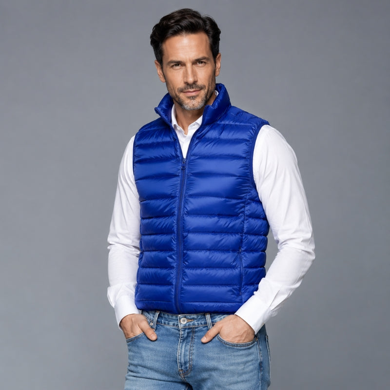 A man wearing a royal blue puffer vest over a white long-sleeve shirt and blue jeans, standing against a solid gray studio background.