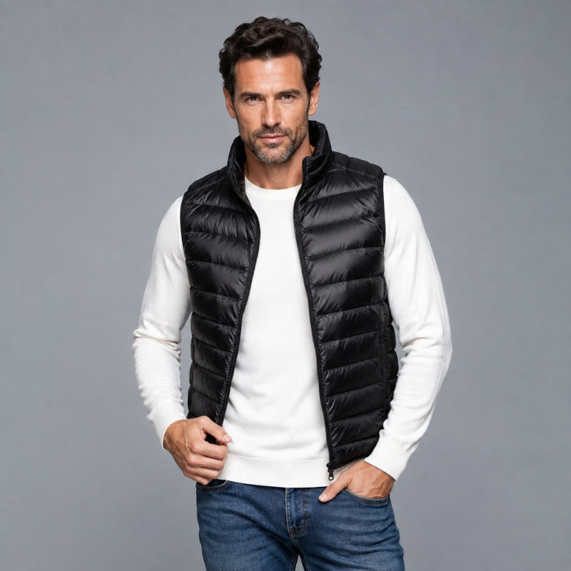 A man wearing a black puffer vest over a white long-sleeve shirt and blue jeans, standing against a solid gray studio background.