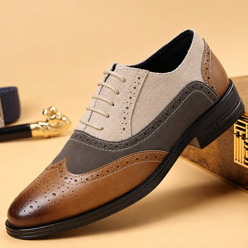 A men's three-tone wingtip oxford shoe with a brown brogue toe, a medium brown suede side panel, and a light beige suede quarter, resting on a gold-toned box against a beige background.