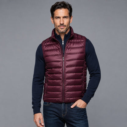 A man wearing a burgundy quilted puffer vest over a navy blue long-sleeve shirt and dark blue jeans, standing against a solid gray studio background.