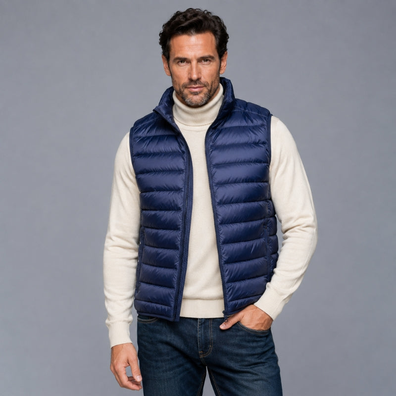 A man wearing a navy blue puffer vest over a cream turtleneck sweater and dark blue jeans, standing against a solid gray studio background.