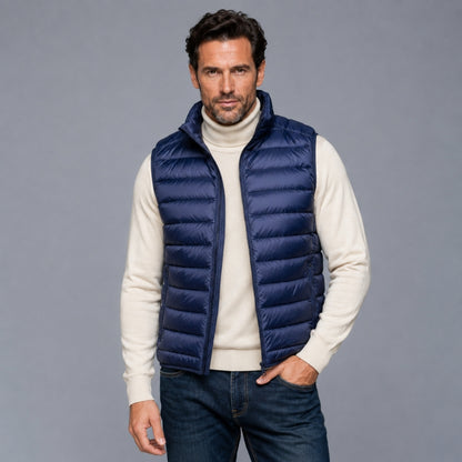 A man wearing a navy blue puffer vest over a cream turtleneck sweater and dark blue jeans, standing against a solid gray studio background.