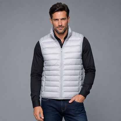 A man wearing a light gray puffer vest over a black long-sleeve shirt and dark blue jeans, standing against a solid gray studio background.