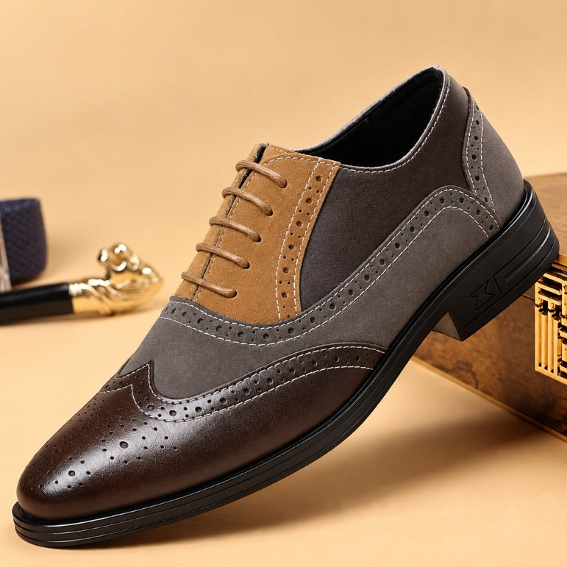 A men's wingtip oxford shoe with a color-blocked design featuring dark brown leather, medium brown suede, and dark gray panels, shown on a beige background with decorative props.