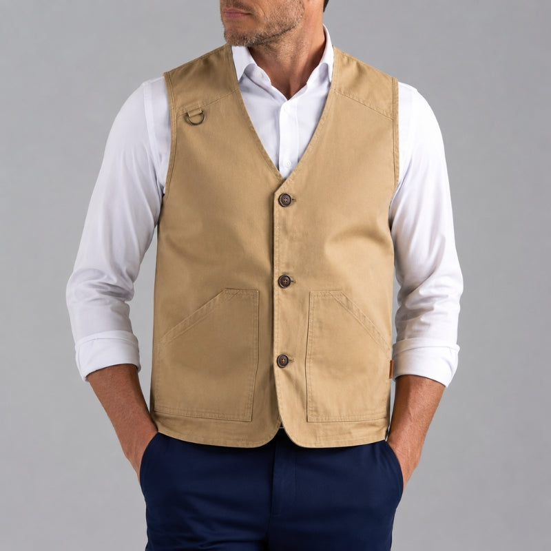 A man wearing a tan utility vest over a white long-sleeve shirt and navy blue trousers, shown from the chin down against a solid gray studio background.