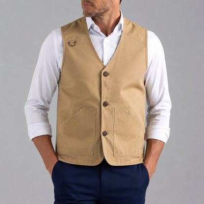A man wearing a tan utility vest over a white long-sleeve shirt and navy blue trousers, shown from the chin down against a solid gray studio background.