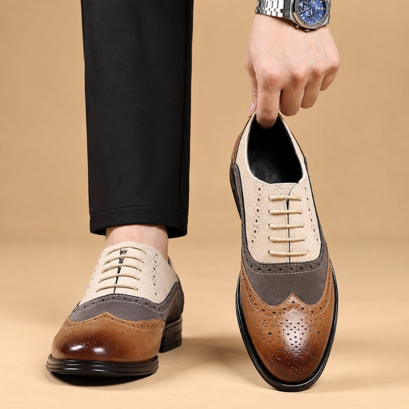 A person wearing a pair of men's three-tone wingtip oxford shoes, with one shoe being held up by a hand. The shoes feature a color-blocked design in brown, beige, and gray, with brogue perforated detailing and laces.